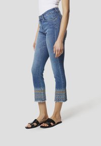 Blauwe spijkerbroek met een cropped zoom, voorzien van ingewikkeld borduurwerk en franjes. Gecombineerd met zwarte platte sandalen.