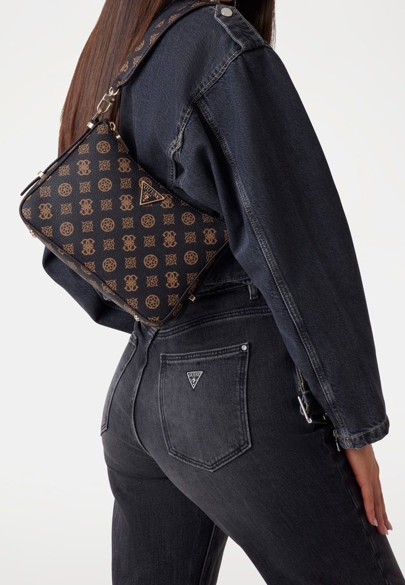 Personne portant une veste en jean noire et un jean avec un sac à bandoulière Guess marron à motifs dans le dos.