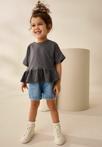 Haut en peplum gris à manches courtes, associé à un short en denim et des baskets montantes blanches. L'arrière-plan présente un objet cylindrique aux teintes neutres.