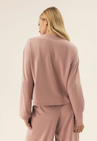 Jasnoróżowy długi rękaw, luźne dopasowanie i okrągły dekolt. Gładka faktura, dół opada na talię, zestawiony z pasującymi spodniami.