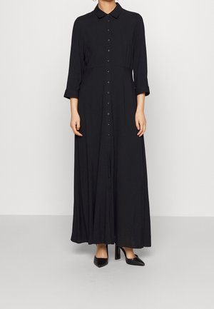 Žena oblečená v dlouhých černých šatech s knoflíky a tříčtvrtečními rukávy, stojící v černých lodičkách s špičatou špičkou na jednobarevném pozadí.