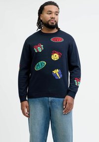Navy sweater with colorful holiday-themed embroidery, featuring gifts, bells, and text. Ribbed cuffs and neckline. Paired with light denim.