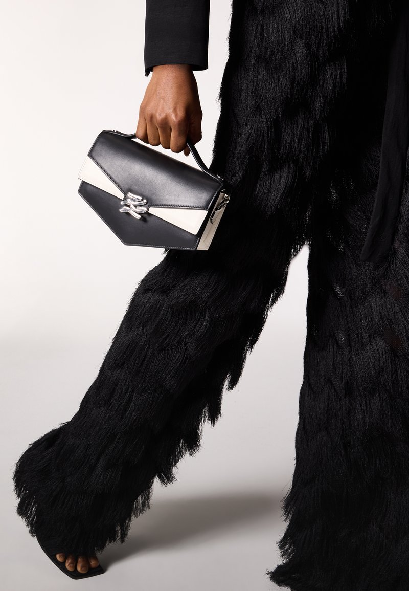 Άτομο που κρατά μια ασπρόμαυρη τσάντα με γωνίες, φοράει μαύρο παντελόνι με φαρδιά πόδια και υφή, και ανοιχτά μαύρα τακούνια.