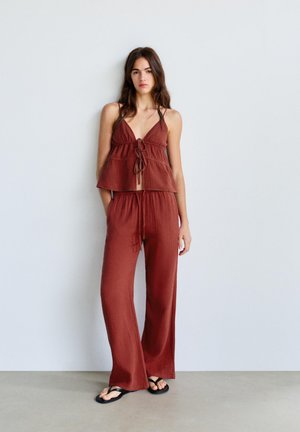 Fiatal nő áll egy fehér falnak támaszkodva, rozsdaszínű ujjatlan felsőt kötőkkel és hozzáillő bőszárú nadrágot, fekete szandállal viselve.