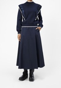 Marineblauwe trui met bloemenborduursel en witte bies, gecombineerd met een bijpassende geplooide rok. G gedragen met zwarte enkellaarsjes.