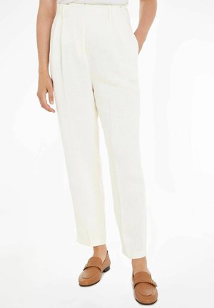 Personne portant un pantalon blanc taille haute avec plis, poches latérales et mocassins en cuir marron sur fond blanc.