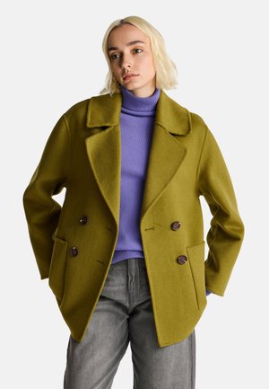 Cappotto di lana doppiopetto verde oliva con due grandi tasche e bottoni marroni, indossato sopra un dolcevita viola e pantaloni grigi.