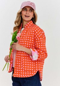 Prešívaná oranžová bunda s ružovým kockovaným vzorom, s ružovým kvetinovým manžetou a dvoma prednými vreckami. Model drží ružové tulipány.