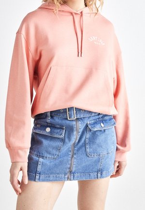 Osoba ubrana w brzoskwiniową bluzę z kapturem i przednią kieszenią oraz niebieską dżinsową mini spódnicę z przednim zamkiem błyskawicznym i dwiema klapkowymi kieszeniami na guziki.