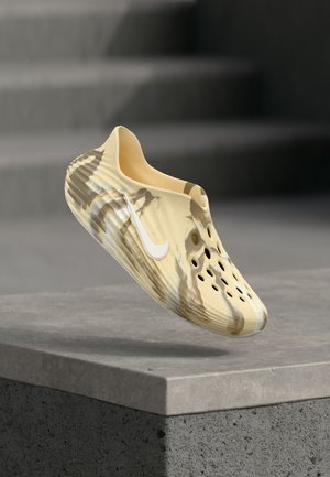 Svetlohnedá a béžová slip-on topánka s ventilačnými otvormi a logom Nike, vznášajúca sa nad sivým betónovým povrchom so rozostreným schodiskom v pozadí.