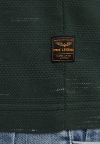 Tela texturizada de color verde oscuro con una etiqueta visible que muestra "PME LEGEND," con texto amarillo y un emblema. Detalle de dobladillo festoneado en la base.