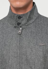 Grå fiskebensjakke med høj krave, to sorte knapper og en sølvfarvet lynlås med et Lacoste-logo broderet i grå.