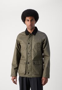 Chaqueta ligera de color verde oliva con cuello de pana negro, botones a presión y dos bolsillos frontales. Presenta una textura suave y un diseño moderno.