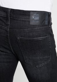 Black denim jeans featuring a leather patch with the logo on the waistband. Single back pocket with curved stitching detail.
