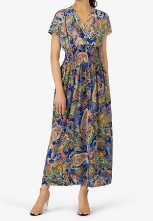 Kobieta ubrana w niebieską maxi sukienkę z wielokolorowymi paisley i kwiatowymi wzorami oraz przezroczyste sandały na kostkę z przezroczystym, masywnym obcasem.