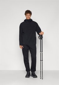Haut de survêtement en polaire noir avec un col montant, associé à un pantalon de survêtement noir et des chaussures noires. Bâtons de trekking légers tenus dans la main droite.