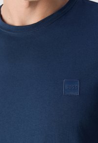 Gros plan d'une personne portant une chemise bleu marine avec un petit logo carré brodé portant la mention "BOSS" sur la poitrine.