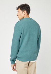Mann mit lockigem Haar, der einen strukturierten türkisfarbenen Pullover und beige Hosen trägt, zur Seite gedreht vor einem einfachen hellen Hintergrund.