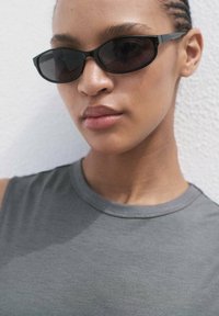 Occhiali da sole rettangolari neri con aste sottili, indossati sopra un top grigio senza maniche, che mostrano un design semplice e elegante con una texture liscia.