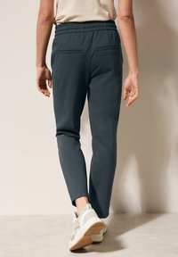 Pantalones jogger de color gris oscuro con una cinturilla elástica y dos bolsillos traseros, combinados con zapatillas de color claro, vistos desde la parte posterior.