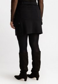 Svart denimskorts med elastisk midja, två bakfickor och en sidficka med knappstängning, bärs över svarta leggings och knähöga stövlar.