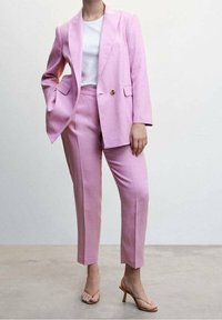 Frau in einem hellrosa maßgeschneiderten Anzug mit einem Blazer mit einem Knopf, einem weißen Oberteil und nudefarbenen Absatzsandalen, die auf einem neutralen Boden steht.