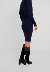 Laivasta sininen neulemekko, jossa on kiinteä siluetti ja pitkät hihat, yhdistettynä mustiin polvihameisiin saappaisiin, joissa on paksu korko ja nahka-akcentteja.