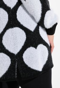 Zwart-witte trui met een fuzzy textuur, grote hartvormen en een geribbelde zwarte zoom. De stof lijkt zacht en comfortabel te zijn.
