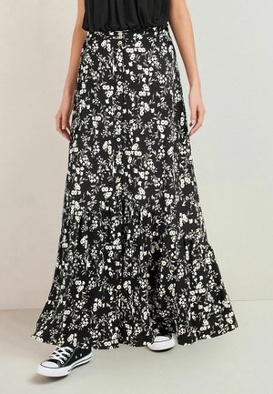 Jupe maxi noire avec imprimé floral blanc et boutons sur le devant, portée avec des baskets noires et blanches et un top noir.