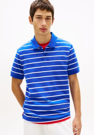 Młody mężczyzna w niebiesko-białej paskowanej koszulce polo na czerwonej koszuli, stojący na jednolitym białym tle.