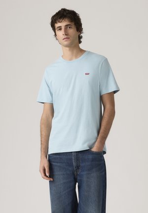 Νέος άνδρας με ανοιχτό μπλε T-shirt Levi's και μπλε τζιν, στέκεται με το ένα χέρι στην τσέπη σε απλό φόντο.