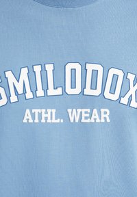 Helles blaues Baumwoll-Sweatshirt mit weißem erhabenem Text "SMILODON ATHL. WEAR", mit Rundhalsausschnitt und geripptem Muster.