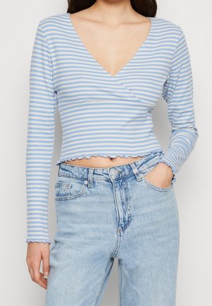 Frau trägt ein langärmeliges, hellblau-weiß gestreiftes V-Ausschnitt-Bauchfreies Oberteil mit Rüschensaum und hochtaillierte hellblaue Jeans.