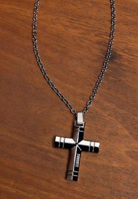 Silverfärgad metalkors hänge med svarta accenter, med en texturerad design, fäst vid ett länkad kedja halsband på en träytan.