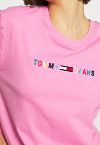 Osoba oblečená v růžové košili s vícebarevným textem "TOMMY JEANS" a na hrudi vyšitým obdélníkovým logem v červené, bílé a černé barvě.