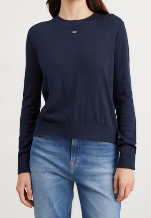 Femme portant un pull à manches longues bleu marine avec un petit logo au niveau de la poitrine et un jean en denim bleu clair.