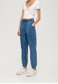Bílé ribbované crop top s výřezem, kombinované s světle modrými denimovými kalhotami typu joggers s elastickými manžetami a postranními kapsami. Bílé tenisky.