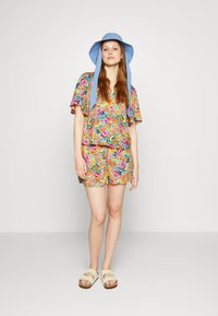 Conjunto colorido com estampado floral, featuring uma blusa larga e calções a condizer, complementado por um chapéu de aba larga azul claro e sandálias beges.