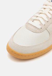 Beige sporta apavi ar gludu kanvasu un zamšiem, apaļo purngali, kontrastējošu gumijas zoli un baltām auklām ar izšuvumiem.
