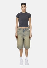 T-shirt aderente grigio scuro abbinato a bermuda in denim azzurro chiaro e sneakers bianche. Il modello si trova di fronte a uno sfondo bianco semplice.