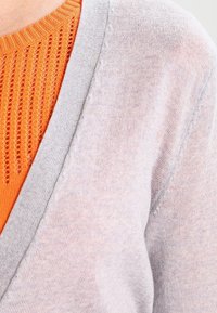 Ljusgrå tröja med V-ringning; lager över en orange stickad topp med öppna stickmönster. Mjuk textur synlig i båda plaggen.