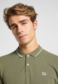 Olive vihreä polo-paita, jossa on kaulus, kaksi nappia ja valkoiset reunat. Vasemmassa rinnassa on pieni kirjailtu "Lee" -logo.