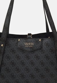 Black textured tote bag featuring a monogram pattern, gold hardware accents, and embossed logo that reads "GUESS EST 1981."