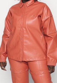 Giacca in similpelle color corallo con chiusura a bottoni a pressione, collo con risvolto e polsini elasticizzati, abbinata a pantaloni coordinati con dettagli a scatto.