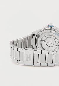 Ezüst rozsdamentes acél óra fém karkötővel, kerek tok, kék korona részlet és látható hátlap, amely az összetett mechanikát mutatja.