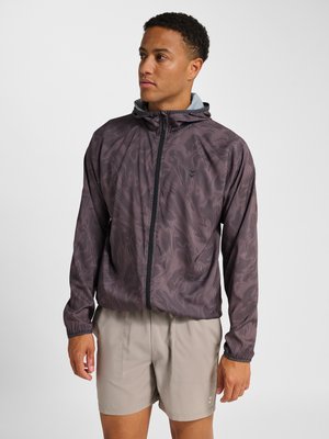 Mann trägt eine braun gemusterte Kapuzenjacke mit Reißverschluss und beige Shorts, steht und schaut nach links vor einem einfarbigen Hintergrund.