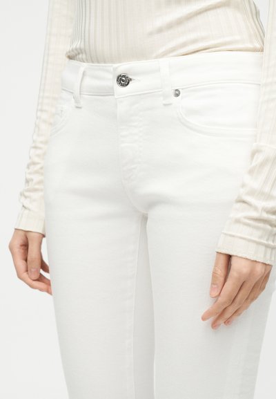 Κοντινό πλάνο ενός ατόμου που φοράει λευκά τζιν με ψηλή μέση και μπροστινές τσέπες, μαζί με μπεζ ριμπ μπλούζα μακρυμάνικη, εστιάζοντας στη μέση και τα χέρια.