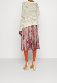 Pull en maille crème avec des mailles ouvertes, associé à une jupe plissée rose à fleurs. Des collants orange et des bottes transparentes à talons complètent le look.