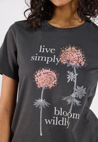 Svart bomullstr�öja med blommiga grafiska tryck i rosa och grått med texten "leva enkelt" och "blomma vilt" i vitt.