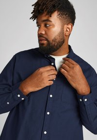 Marineblaues Baumwollhemd mit langen Ärmeln, weißen Knöpfen und einer Brusttasche. Mann richtet den Kragen. Darunter ein weißes Rundhals-T-Shirt.